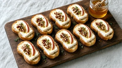 Wooden board of about ten apple honey Brie crostini with red apple slices, chopped pecans, thyme, and a honey jar with dipper