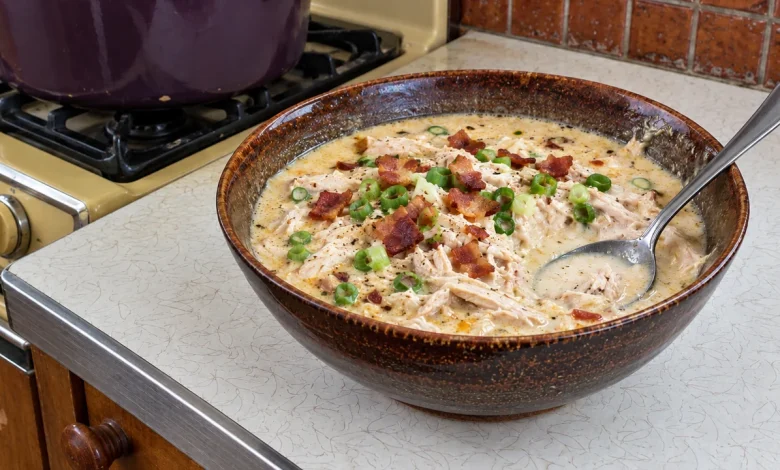 A bowl of creamy crack chicken soup with plenty of shredded chicken, crisp bacon pieces and sliced green onions, a spoon resting in the bowl