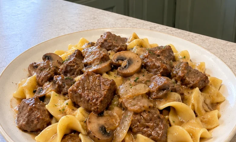 Dutch oven of homemade beef and noodles style stew with browned steak cubes, mushrooms and medium-brown gravy beside buttered wide egg noodles